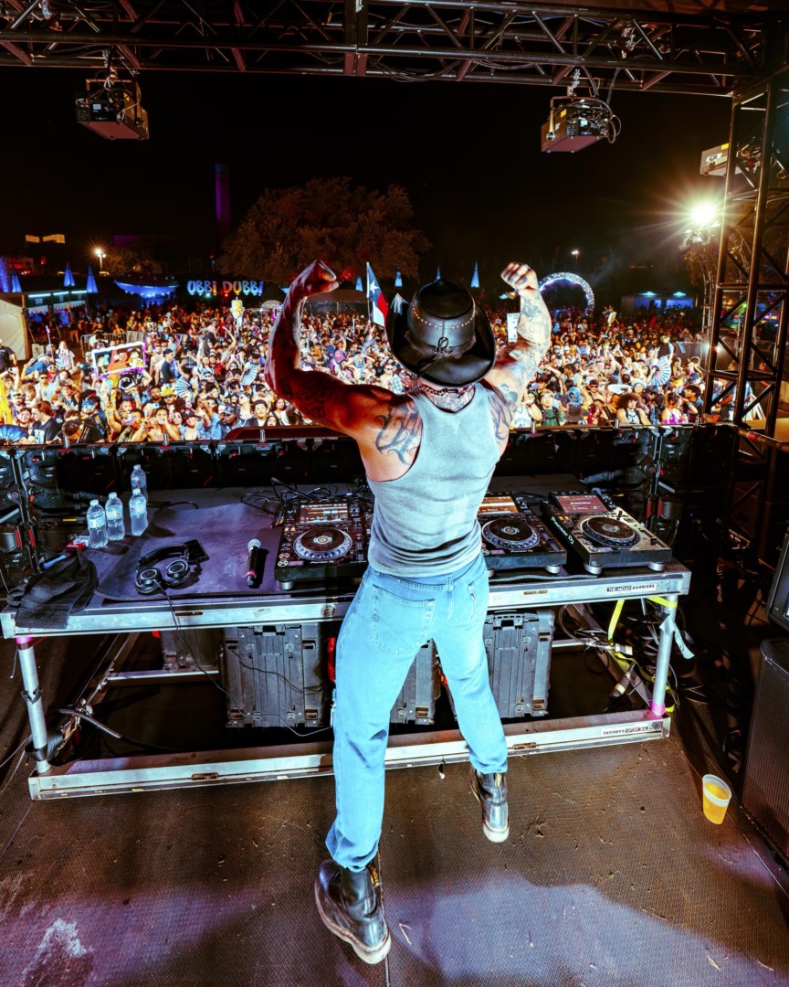 Lil Texas flexing his guns in the air behind the turn tables