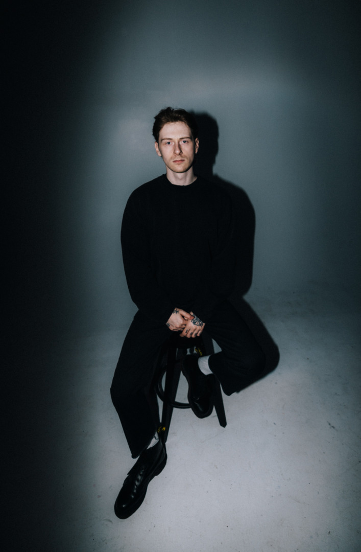 Tisoki psoing in all black seated upon a chair or stool